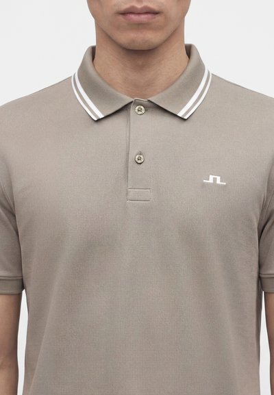 Homme portant un polo taupe avec des doubles rayures blanches sur le col et un petit logo blanc sur la poitrine gauche.