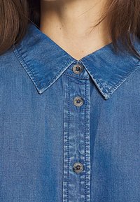 Camicia in denim realizzata in un tessuto blu chiaro, con colletto, cinque bottoni sulla parte anteriore e dettagli di cucitura visibili.