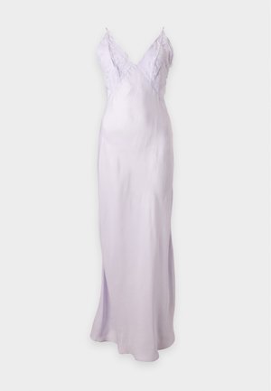 Femme portant une robe nuisette en satin lavande clair avec des détails en dentelle, debout avec une main sur la hanche et des escarpins blancs à bout pointu sur fond uni.