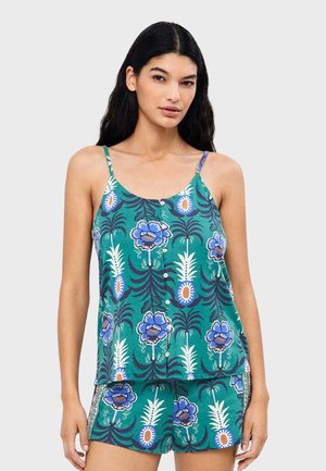 Mujer con camisola verde y pantalones cortos con patrones florales y de piñas azules y blancos, posando contra un fondo gris liso.