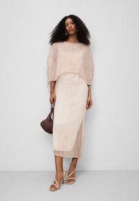 Falten Midi-Kleid in hellem Beige mit einer kurzen Überlage. Verfügt über transparente Textur, subtile Schimmerung und einen seitlichen Schlitz. Kombiniert mit Riemchen-Heels und einer geflochtenen Tasche.