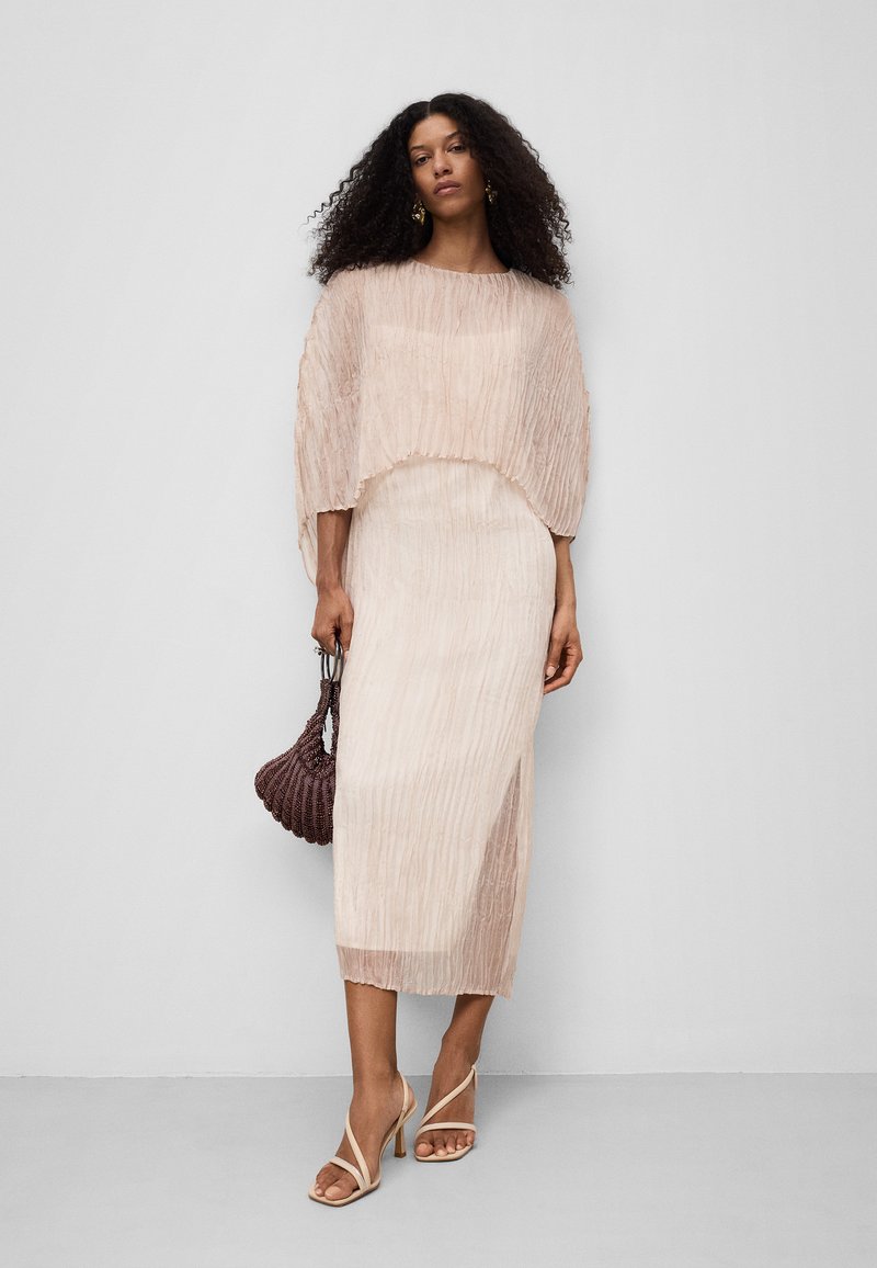 Falten Midi-Kleid in hellem Beige mit einer kurzen Überlage. Verfügt über transparente Textur, subtile Schimmerung und einen seitlichen Schlitz. Kombiniert mit Riemchen-Heels und einer geflochtenen Tasche.