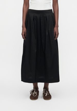 Jupe plissée noire mi-longue portée avec des chaussures à lacets marron et un top blanc sans manches, sur un fond clair uni.