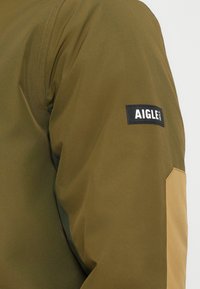 Veste vert olive avec une finition texturée. Présente un panneau beige sur l'épaule et une étiquette cousue sur le bras avec le logo "AIGLE".