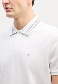 Bărbat purtând tricou polo alb cu dungi duble gri pe guler și mic logo brodat "cK" pe piept, decupat la gât și umăr.