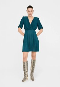 Sinakas rohelise tooniga plisseeritud kleit, millel on sügav V-kaelus ja punn-säärega varrukad. Sobitatud põlveni ulatuvate, metalliklikus madu-mustriga saapadega.