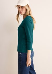 Tyrkysový přiléhavý top s dlouhými rukávy a výstřihem do V s rukávy po třičtvrtě, zkombinovaný s klasickými modrými džínami. Modelka má na sobě bílou kšiltovku.