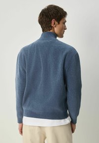 Mėlynas megztinis su aukštu apykakle, tekstūruotu audiniu ir tampriu hemeliu. Rodytas iš nugaros, uždėtas ant balto marškinėlio.