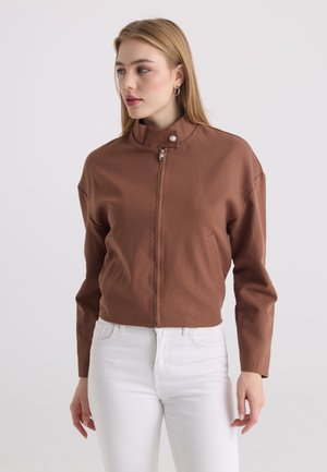 Jeune femme portant une veste marron zippée avec un col à pression et un pantalon blanc, regardant vers sa gauche sur un fond uni.