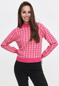 Różowy sweter z dzianiny z wzorem houndstooth, z prążkowanym kołnierzem i mankietami oraz dopasowanym krojem. Nosi się go z czarnymi spodniami.