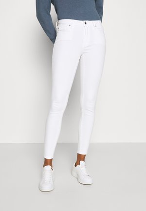 Persoon die witte skinny jeans, witte sneakers en een blauw top met lange mouwen draagt, staand tegen een eenvoudige lichte achtergrond.