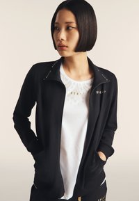 Giacca nera con zip e tasche laterali; presenta lettere dorate sul colletto e sul petto. T-shirt bianca sotto con dettaglio a motivo sottile.