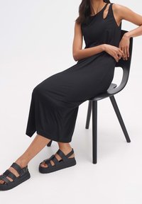 Robe noire sans manches avec une découpe unique à l'épaule, texture lisse et longueur jusqu'aux chevilles. Associée à des sandales noires épaisses.