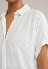 Camicia bianca a maniche corte con tessuto testurizzato, colletto con bottoni e sottili righe verticali. Presenta una placca frontale e vestibilità comoda.