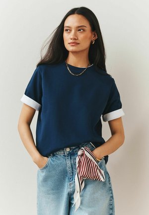 Junge Frau mit langen dunklen Haaren, die ein marineblaues Hemd mit hochgekrempelten Ärmeln, hellblaue Jeans, eine goldene Halskette trägt und einen gestreiften Schal hält.