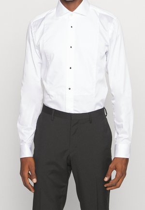 Mand iført en hvid smoking-skjorte med sorte knapper og sorte bukser, stående med armene afslappet ned langs siderne.