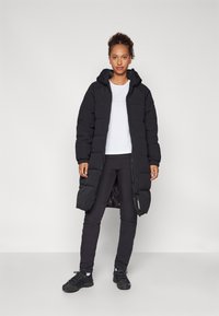 Lang sort polstret frakke med hætte, med en frontlynlås og elastiske manchetter, parret med sorte leggings og sneakers, på en lys baggrund.
