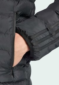 Detailný záber ruky, ktorá sa vkladá do zipsového vrecka čiernej prešívanej bundy so skladaným dizajnom rukávov.