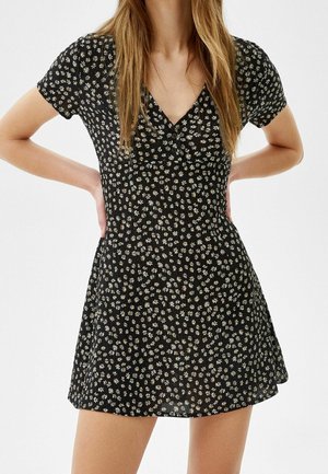 Woman wearing a short black dress with white daisy floral print, short sleeves, and a V-neckline with buttons.