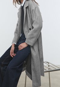 Grijze wollen trenchcoat met grote revers en twee knopen, gecombineerd met donkere jeans. Beide kledingstukken hebben een relaxte pasvorm en zichtbare naden.