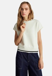 Bílý krátký rukáv kroptop s kulatým výstřihem, který má černobílý pruhovaný spodní lem. Modelka má na sobě tmavé denimové kalhoty.