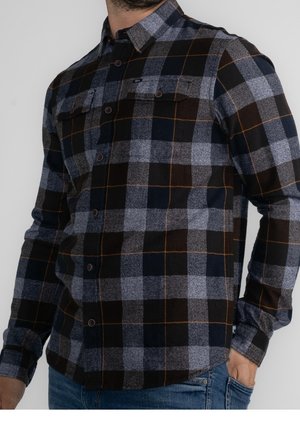 Homme portant une chemise à carreaux bleu foncé, grise et noire à boutons, avec des poches poitrine, et un jean bleu, main dans la poche.