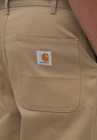 Pantaloni bež boje napravljeni od čvrste tkanine, s stražnjim džepom na kojem se nalazi bijela oznaka s narančastim logom. Vidljive šavove.