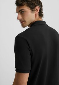 Polo nero con colletto classico, maniche corte e tessuto texturizzato. Il modello guarda oltre la spalla, mostrano la vestibilità della camicia.