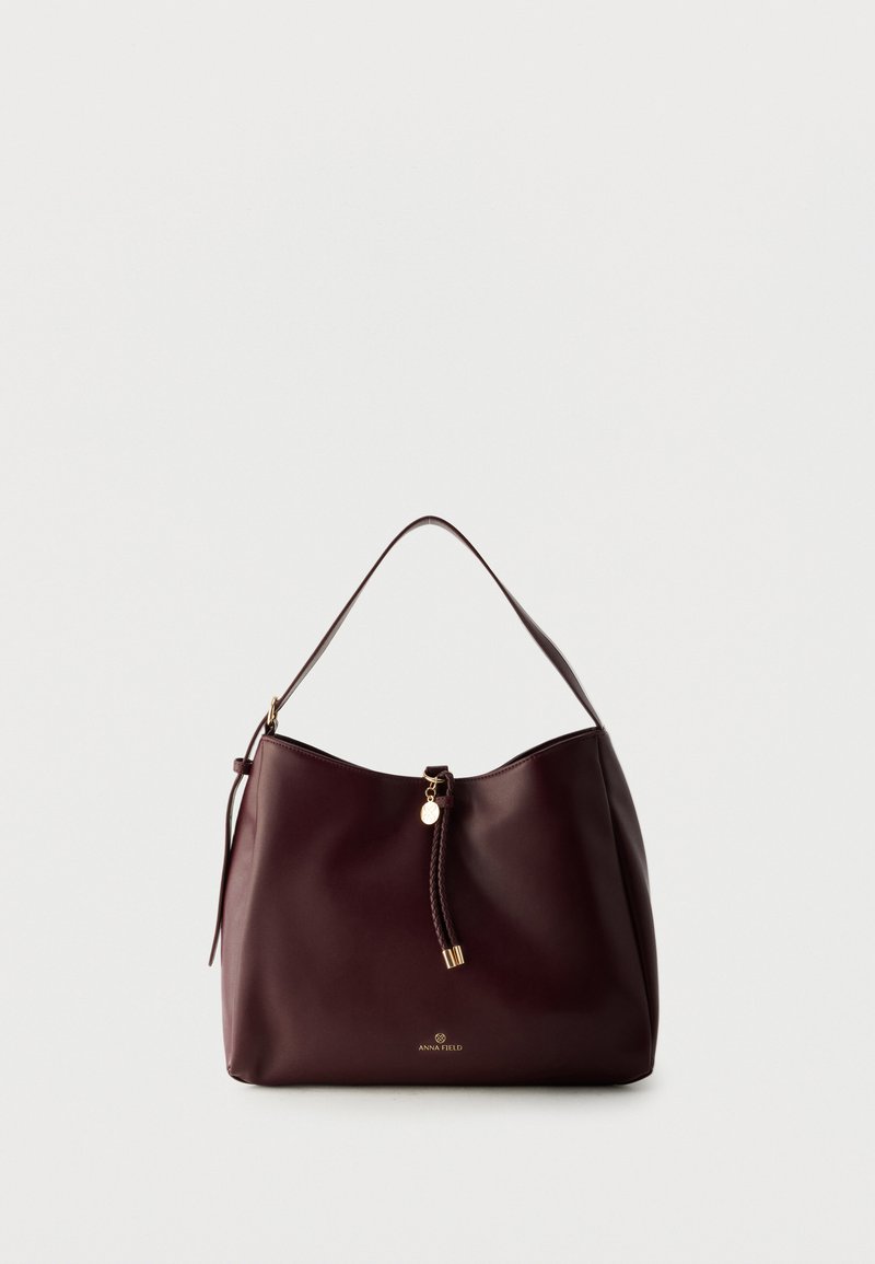 Sac à épaule en cuir marron foncé avec un pompon tressé, des accents dorés et le logo Anna Field à l'avant, sur un fond clair uni.