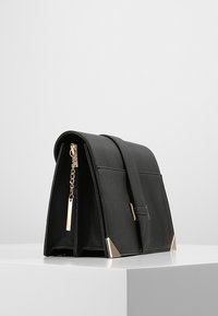 Sac à main en cuir noir avec un design structuré, comportant un couvercle à rabat, des accents en métal doré et un détail en chaîne sur le côté.
