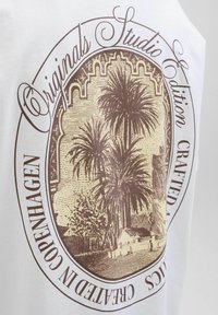 Hvid stof t-shirt med et brunt grafisk tryk af palmer og en bygning, omgivet af dekorativ tekst i en cirkulær opsætning.