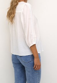 Blouse blanche à manches longues avec une coupe décontractée et une texture subtile ; présente des coutures décoratives le long de la couture d'épaule. Associée à un jean bleu.