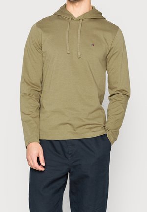 Man draagt een olijfgroene hoodie met lange mouwen, voorste trekkoorden en een klein logo op de borst, gecombineerd met donkerblauwe broek.
