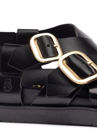 Schwarze Leder-Sandale mit auffälligen goldenen Schnallen, strukturierten Riemen und einer glatten Sohle. Detail des emblemierten Logos an der Seite.