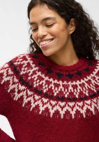 Pull en tricot rouge avec un col rond, arborant un motif décoratif en blanc et noir à motifs géométriques. Texture douce.