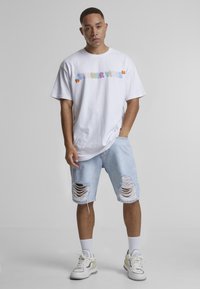 Hvid oversized t-shirt med farverig "SUMMER VIBES" grafik, sammen med lyseblå revnede denim shorts og hvide sneakers.