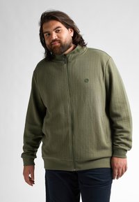 Chaqueta de cremallera color verde oliva con un patrón texturizado, cuello alto y cremallera frontal. Presenta puños y dobladillo de punto acanalado, acompañada de un pequeño logo.