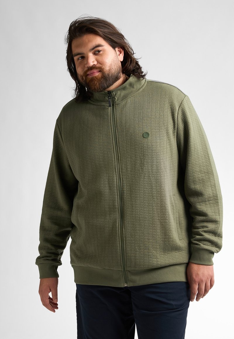 Chaqueta de cremallera color verde oliva con un patrón texturizado, cuello alto y cremallera frontal. Presenta puños y dobladillo de punto acanalado, acompañada de un pequeño logo.