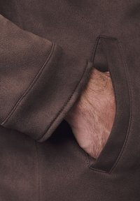 Rękaw brązowej zamszowej kurtki z kieszenią naszywaną, w której częściowo widoczna jest ręka. Miękka tekstura i schludne detale przeszycia.