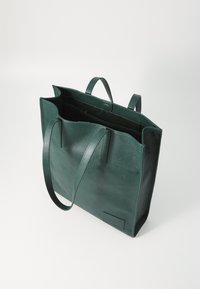 Vihreä nahkatote laukku, jossa on tukevat kahvat, teksturoitu pinta, avoin yläpään ja pieni sisätasku. Sivulla on brändätty logopatch.