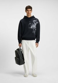 Felpa navy con una grafica astratta bianca, abbinata a pantaloni bianchi e scarpe grigie. Zaino nero testurizzato tenuto in una mano.