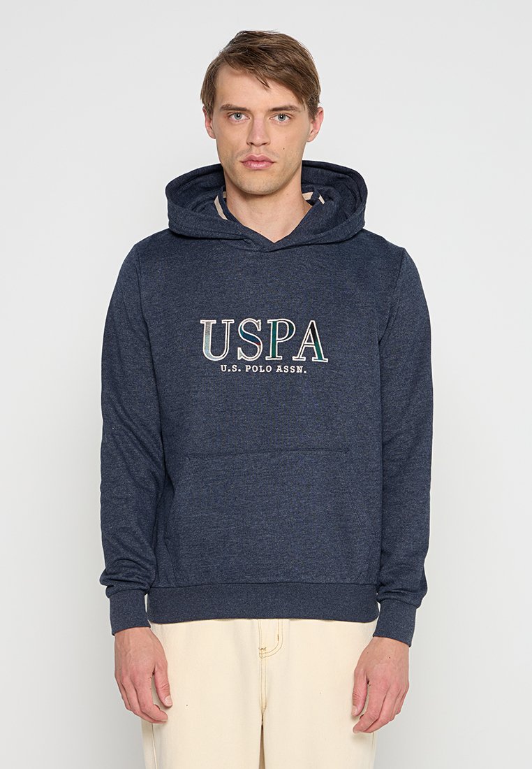 Jeune homme portant un hoodie bleu marine de U.S. Polo Assn. et un pantalon beige clair, se tenant devant un fond blanc uni.