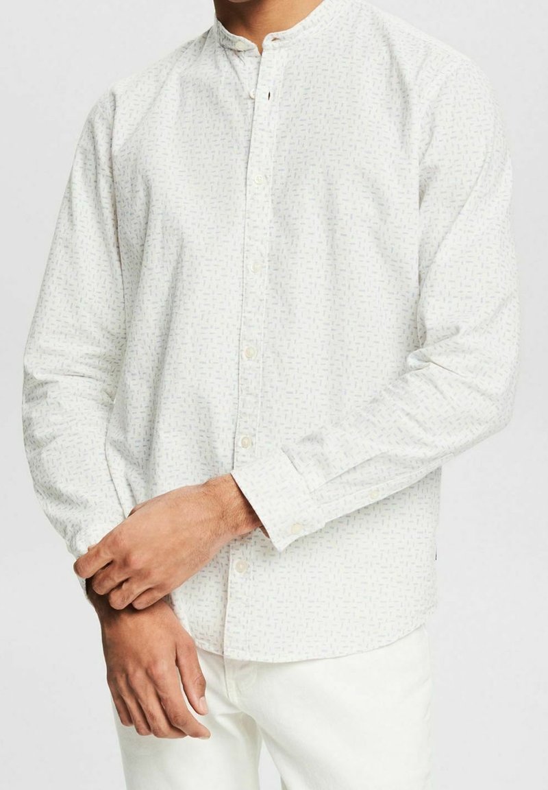 Homme portant une chemise blanche à manches longues avec un motif subtil et un pantalon blanc, ajustant sa manche gauche avec sa main droite.