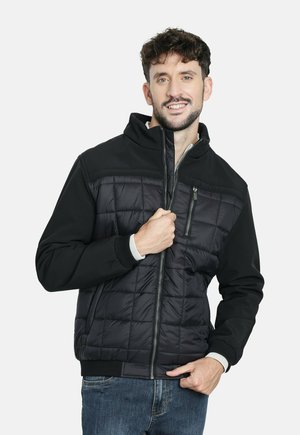 Hombre sonriendo, vistiendo una chaqueta negra acolchada con bolsillos con cremallera y jeans azules, de pie contra un fondo blanco liso.