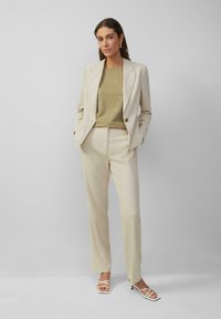 Costume beige ajusté avec un blazer à un bouton et un pantalon droit, associé à un T-shirt graphique olive clair et des talons blancs à lanières.