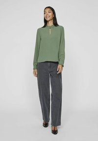 Grüne Bluse mit Rüschensaum und Schlüssellochdetail, lange Ärmel. Kombiniert mit hoch taillierten dunklen Jeans und spitzen Schuhen.