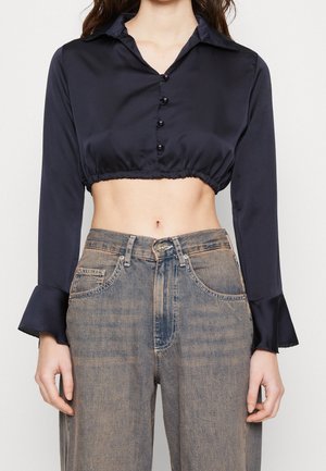 Femme portant un chemisier en satin noir court à boutons avec des poignets évasés et un jean bleu délavé taille haute.