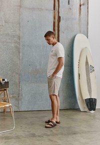 Homme en t-shirt blanc, short beige et sandales, debout à l'intérieur près de deux planches de surf appuyées contre un mur en métal.