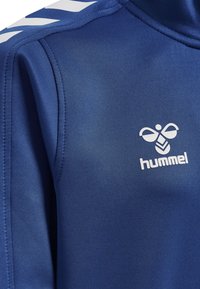 Niebieska kurtka sportowa wykonana z gładkiego materiału, z białymi paskami w kształcie chevron na ramionach i białym logo na klatce piersiowej.