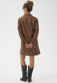 Abito in pelle marrone con frange lungo le maniche, abbinato a stivali cowboy neri, visto da dietro su uno sfondo neutro.